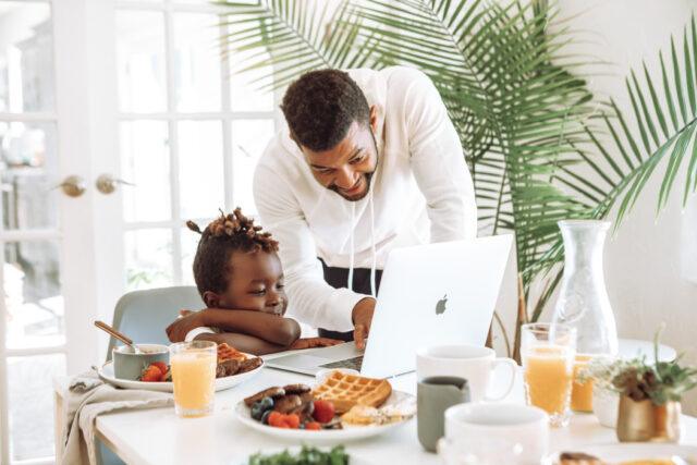 A man and a child at a breakfast table filled with food looking at a laptop